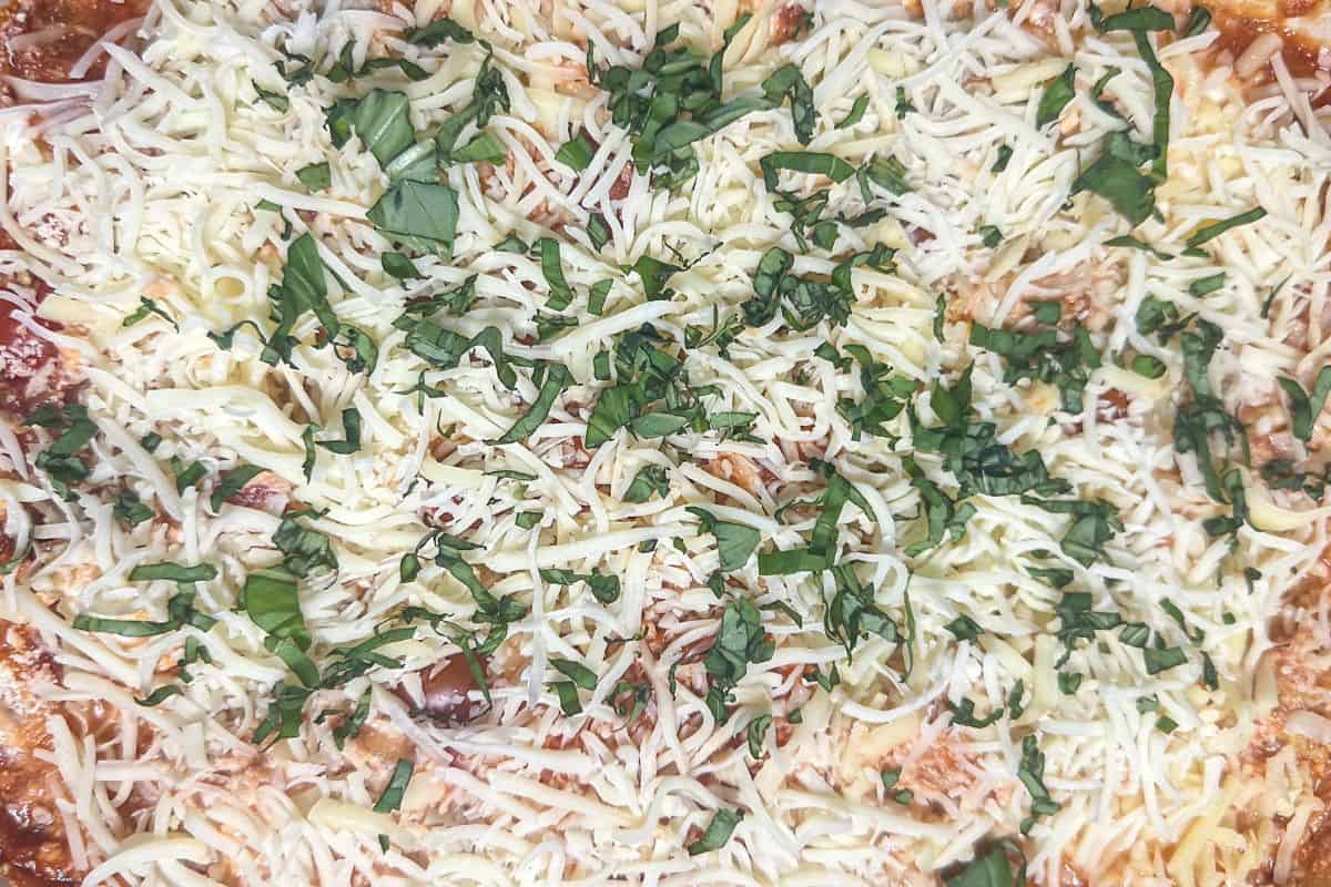 The final layer of spaghetti baked in a casserole dish with shredded cheese and chopped basil spread evenly on top before baking.