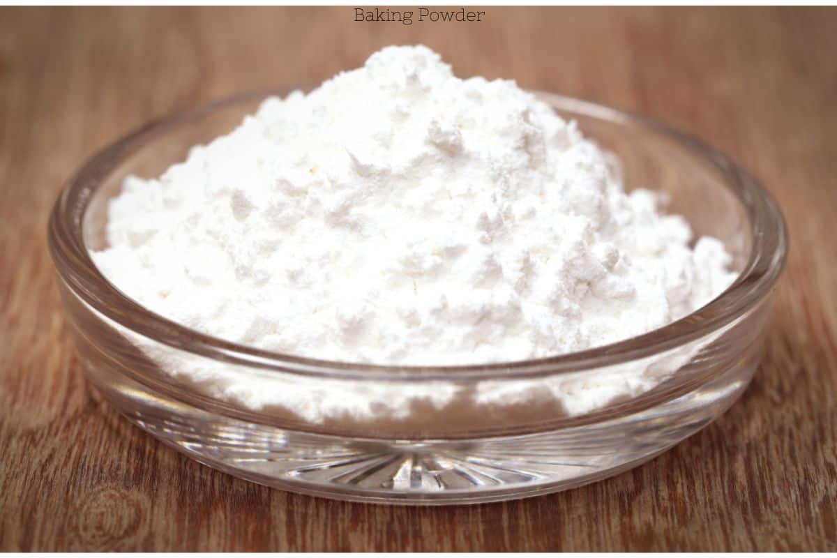 A clear glass bowl filled with white baking powder on a wooden surface.