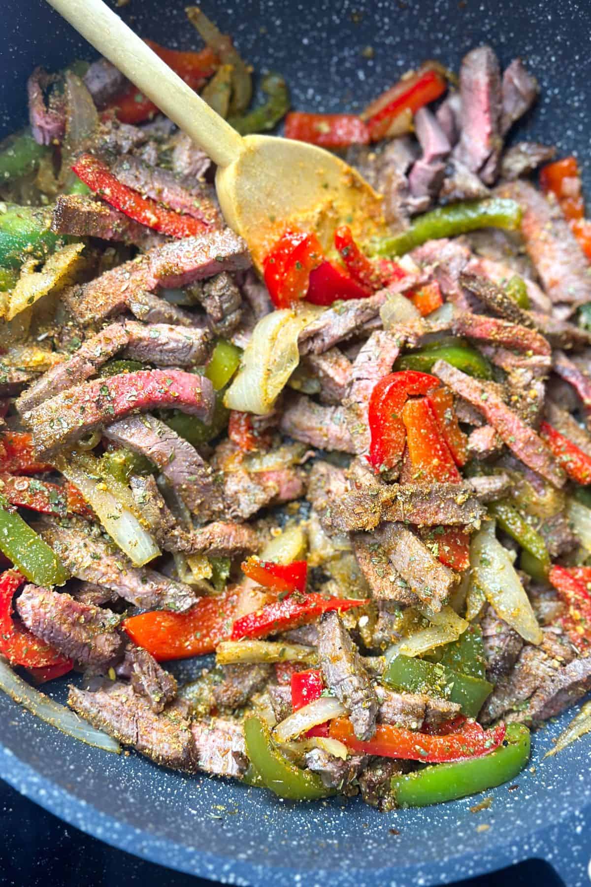 Seasonings being sprinkled over sliced steak and sautéed peppers and onions in a skillet, preparing for the perfect fajita mix.