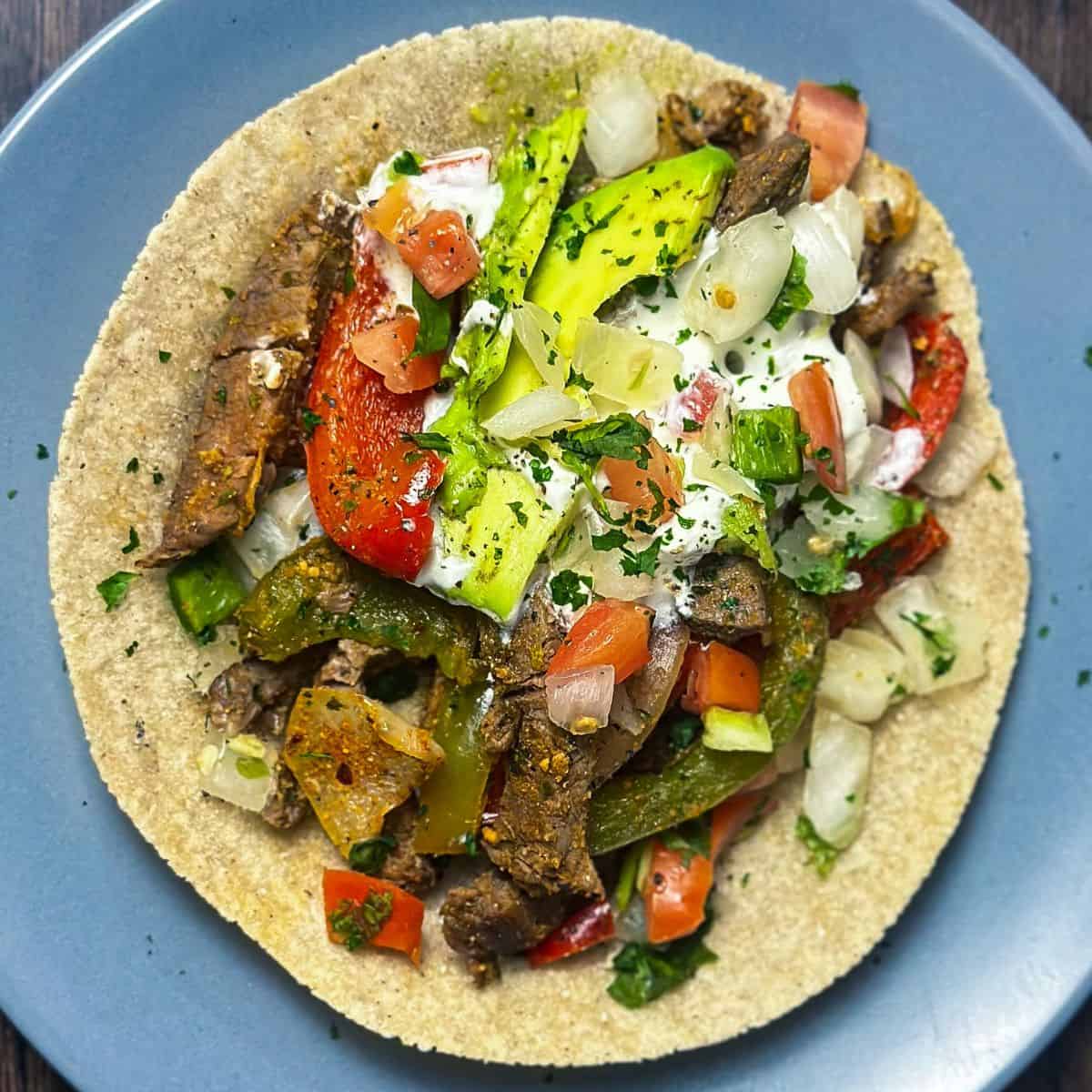 A gluten-free tortilla loaded with seasoned steak, sautéed peppers, onions, and topped with fresh pico de gallo and avocado slices.