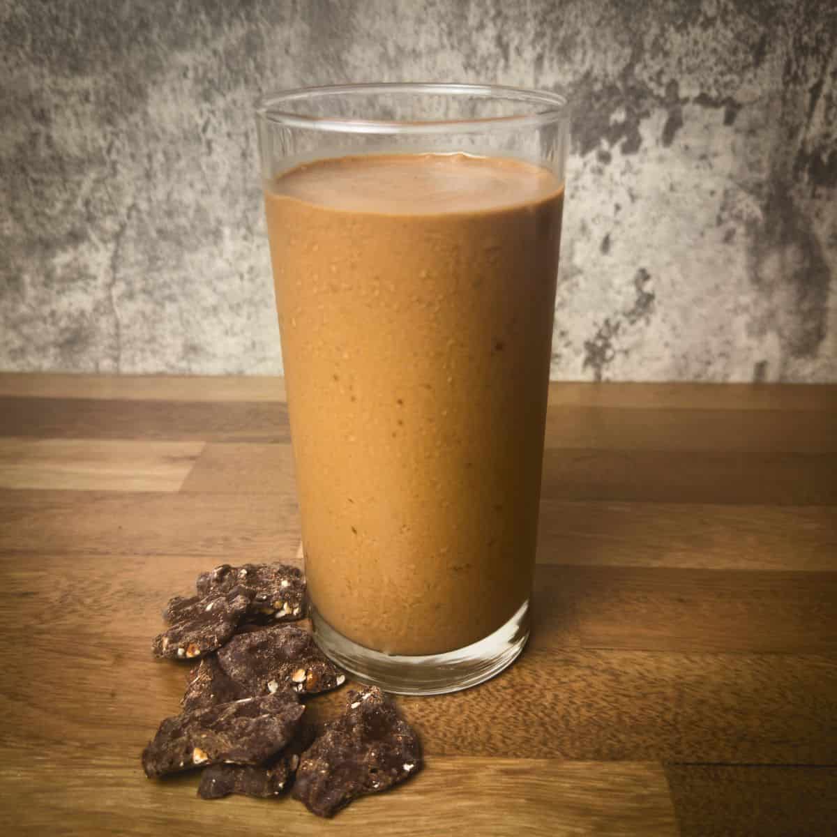 A freshly blended Tropical Smoothie Peanut Butter Cup served in a clear glass, accompanied by chocolate peanut clusters, against a rustic grey background.