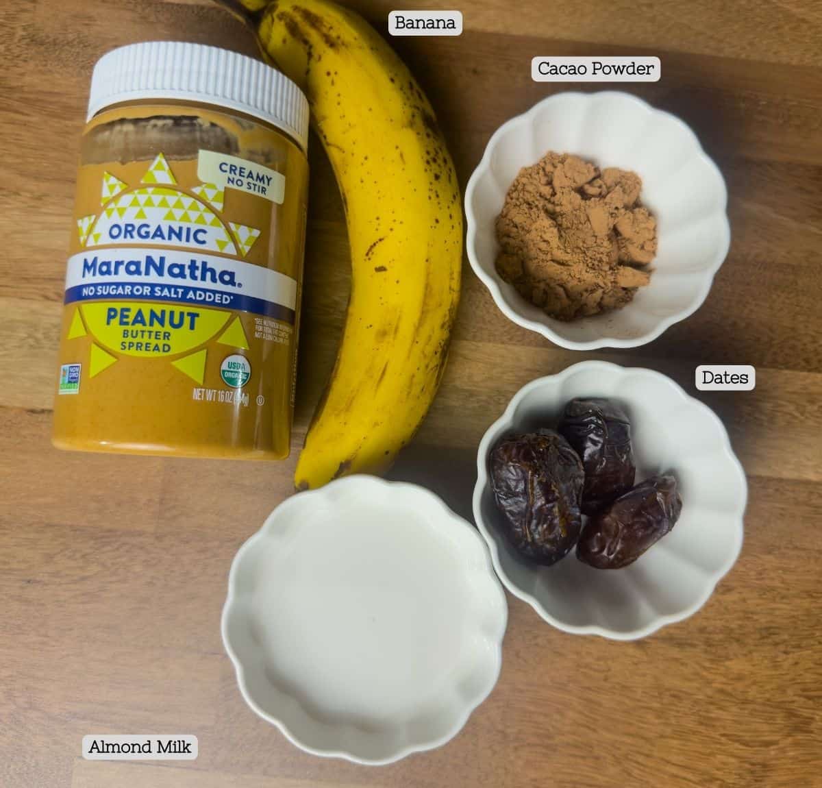 Ingredients for a Tropical Smoothie Peanut Butter Cup neatly laid out, including a banana, cacao powder, Medjool dates, and almond milk with peanut butter in the backdrop.