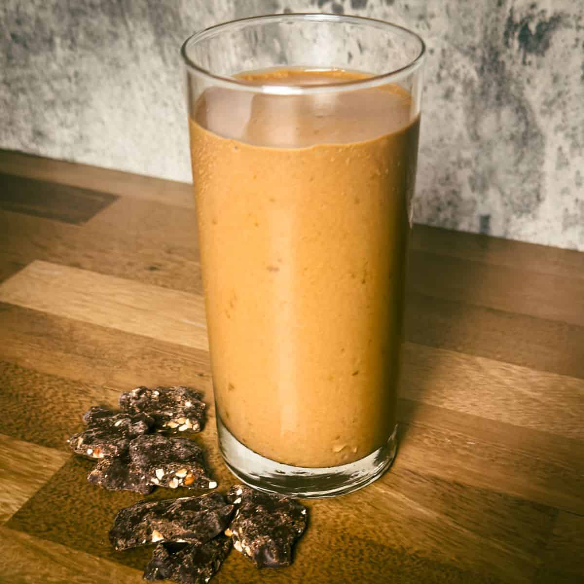 he completed Tropical Smoothie Peanut Butter Cup in a clear glass, with a creamy texture and chocolate peanut clusters to the side, on a wooden tabletop.