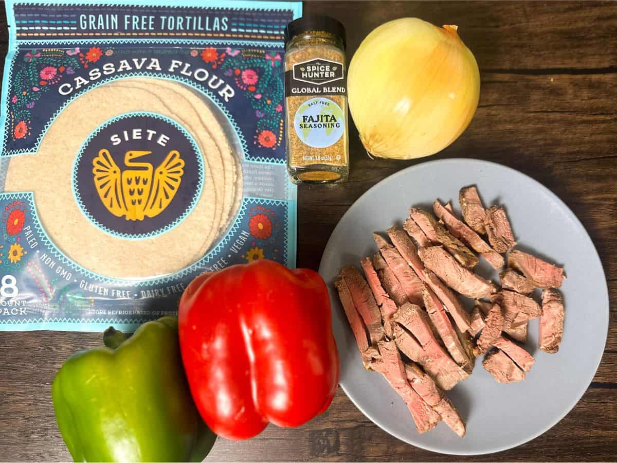 Ingredients for fajitas arranged on a wooden surface, featuring a package of cassava flour tortillas, a bottle of fajita seasoning, fresh bell peppers, onion, and sliced steak.