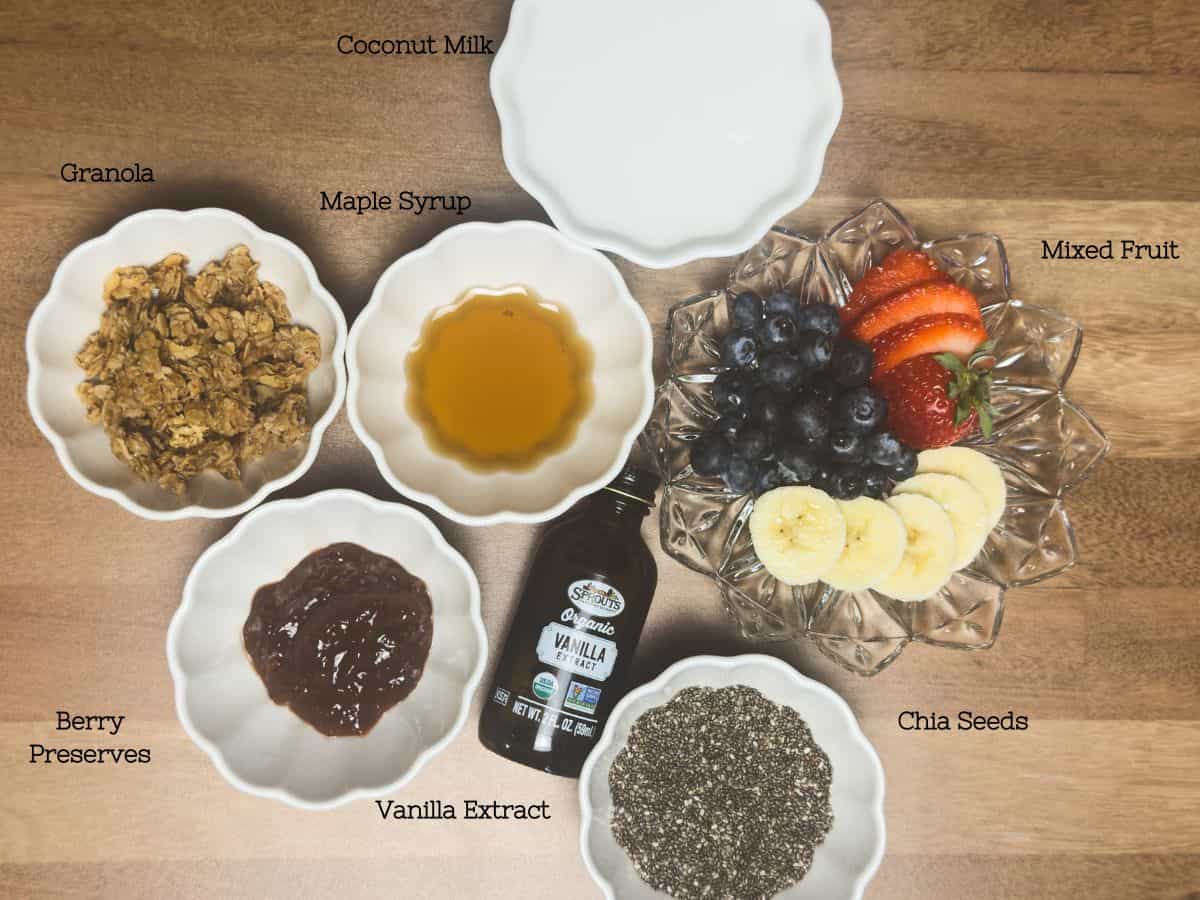 An array of ingredients laid out for making chia seed pudding, including bowls of granola, maple syrup, berry preserves, vanilla extract, mixed fruit, and chia seeds, labeled for clarity.