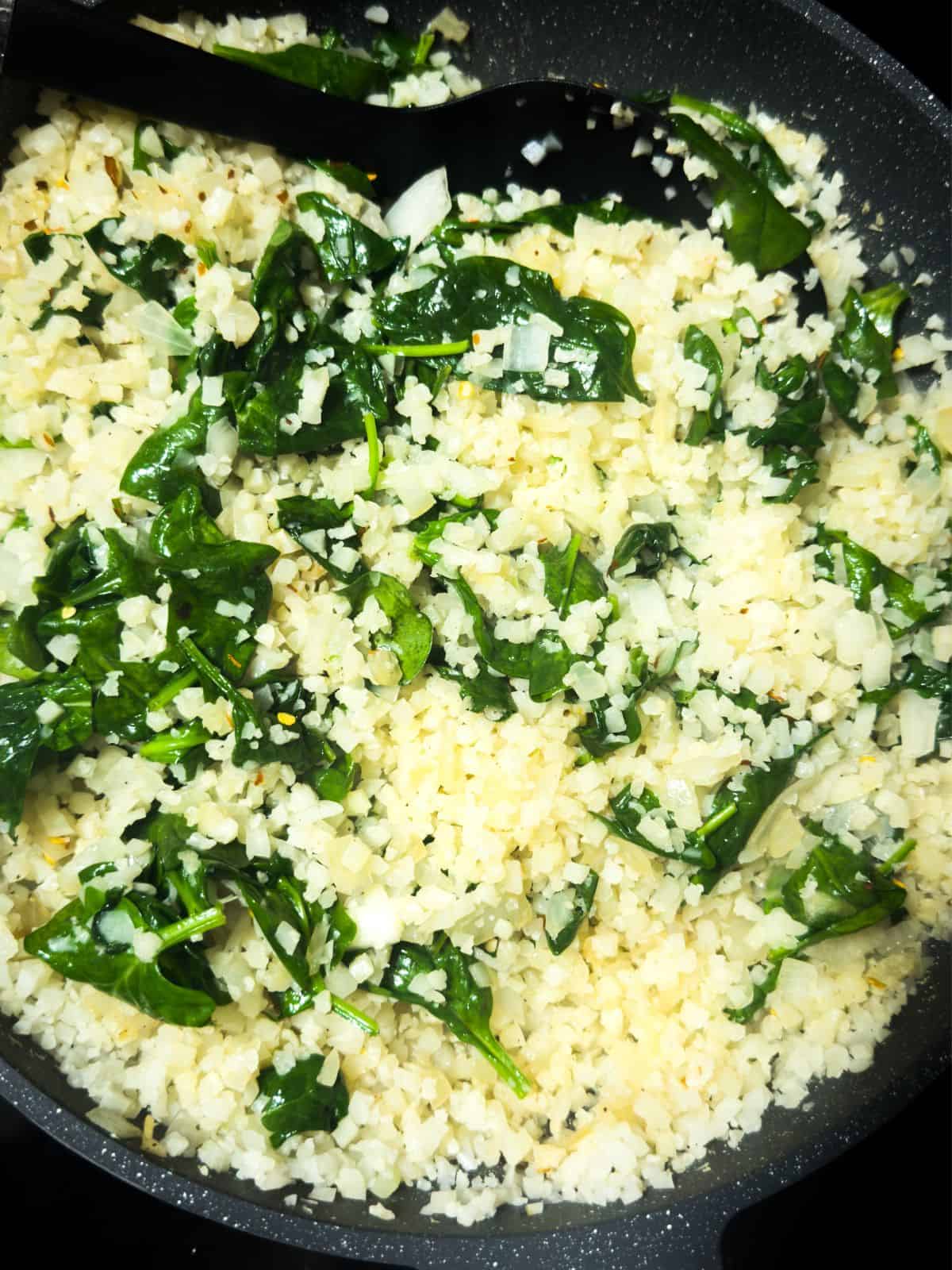 Cauliflower rice being sautéed with spinach in a large skillet for a keto chicken casserole.