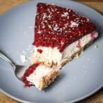 A slice of no-bake berry cheesecake on a grey plate, topped with red berry compote and coconut shavings, with a fork taking a bite.