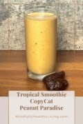 A tall glass of creamy peanut butter smoothie with a golden hue, garnished with two Medjool dates on a wooden table, labeled 'Tropical Smoothie CopyCat Peanut Paradise' from MindfullyHealthyLiving.com