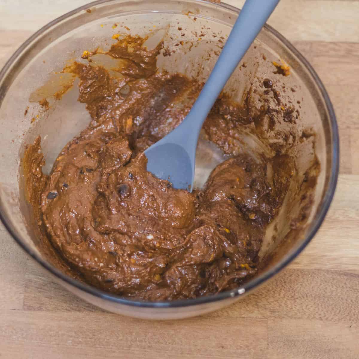 A bowl of thick, chocolate brownie batter with a silicone spatula, indicating the step where dark chocolate chips are folded into the mixture.