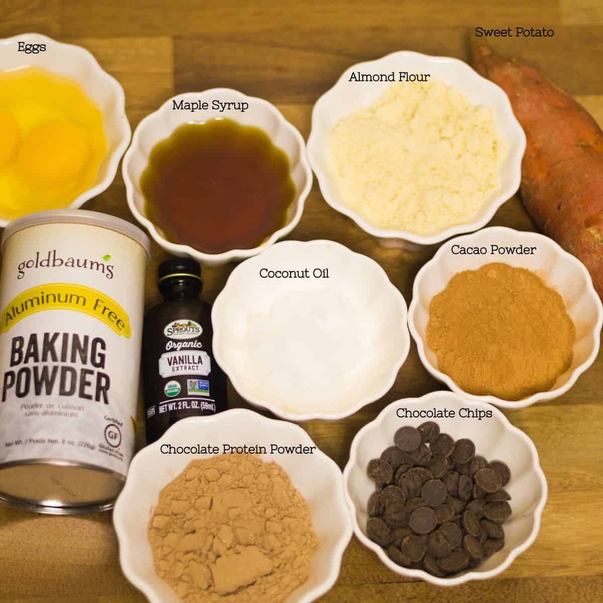 Individual bowls of ingredients laid out for sweet potato protein brownies, including eggs, maple syrup, almond flour, sweet potato, cacao powder, coconut oil, chocolate protein powder, and chocolate chips, ready for mixing.
