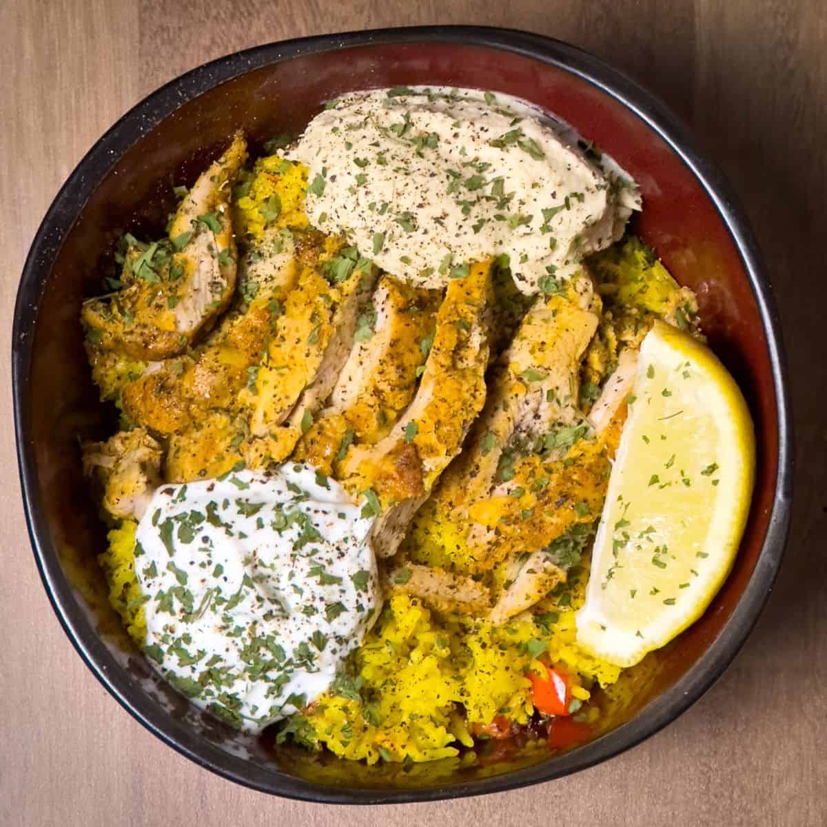 A hearty bowl filled with succulent slices of Chicken Shawarma, nestled on golden Saffron Rice, and topped with a dollop of Tzatziki sauce, accompanied by a fresh lemon wedge.
