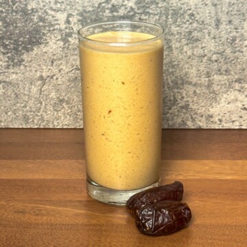 A single serving of peanut butter paradise smoothie presented in a clear glass, with a creamy texture and a couple of Medjool dates beside it on a wooden table.