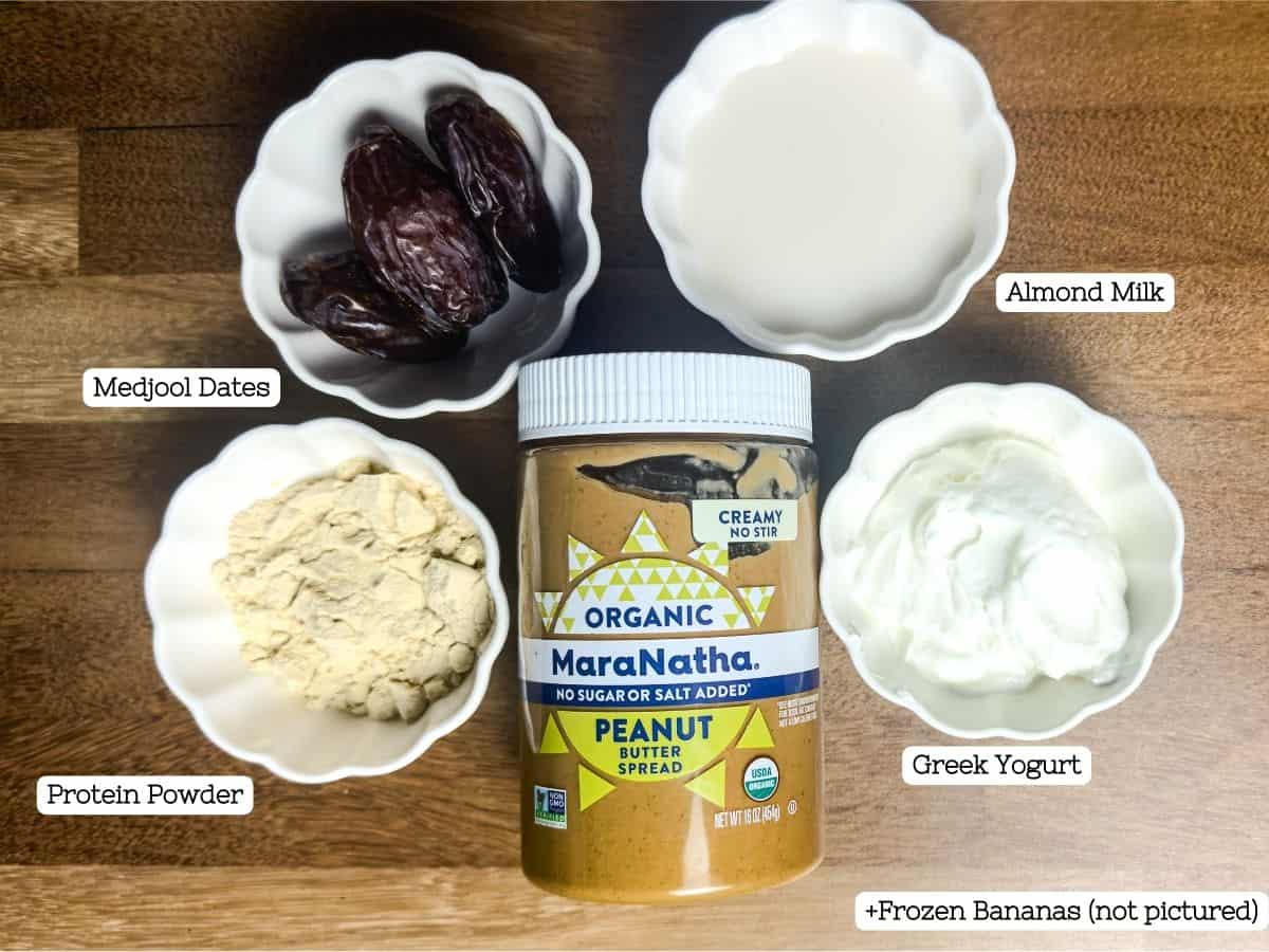 An assortment of ingredients laid out on a wooden surface for a peanut butter paradise smoothie, including Medjool dates, protein powder, Greek yogurt, peanut butter, and almond milk, with a note that frozen bananas are not pictured.