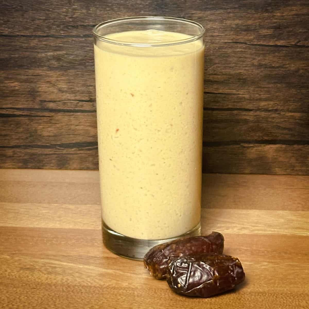 A creamy, blended peanut butter smoothie in a clear glass, exhibiting a rich golden color, placed next to two dark Medjool dates on a wooden tabletop, indicative of the Peanut Paradise flavor.