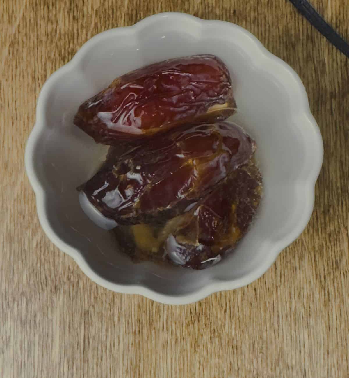 A close-up of a white bowl filled with plump Medjool dates soaking in water to soften, set on a wooden background.
