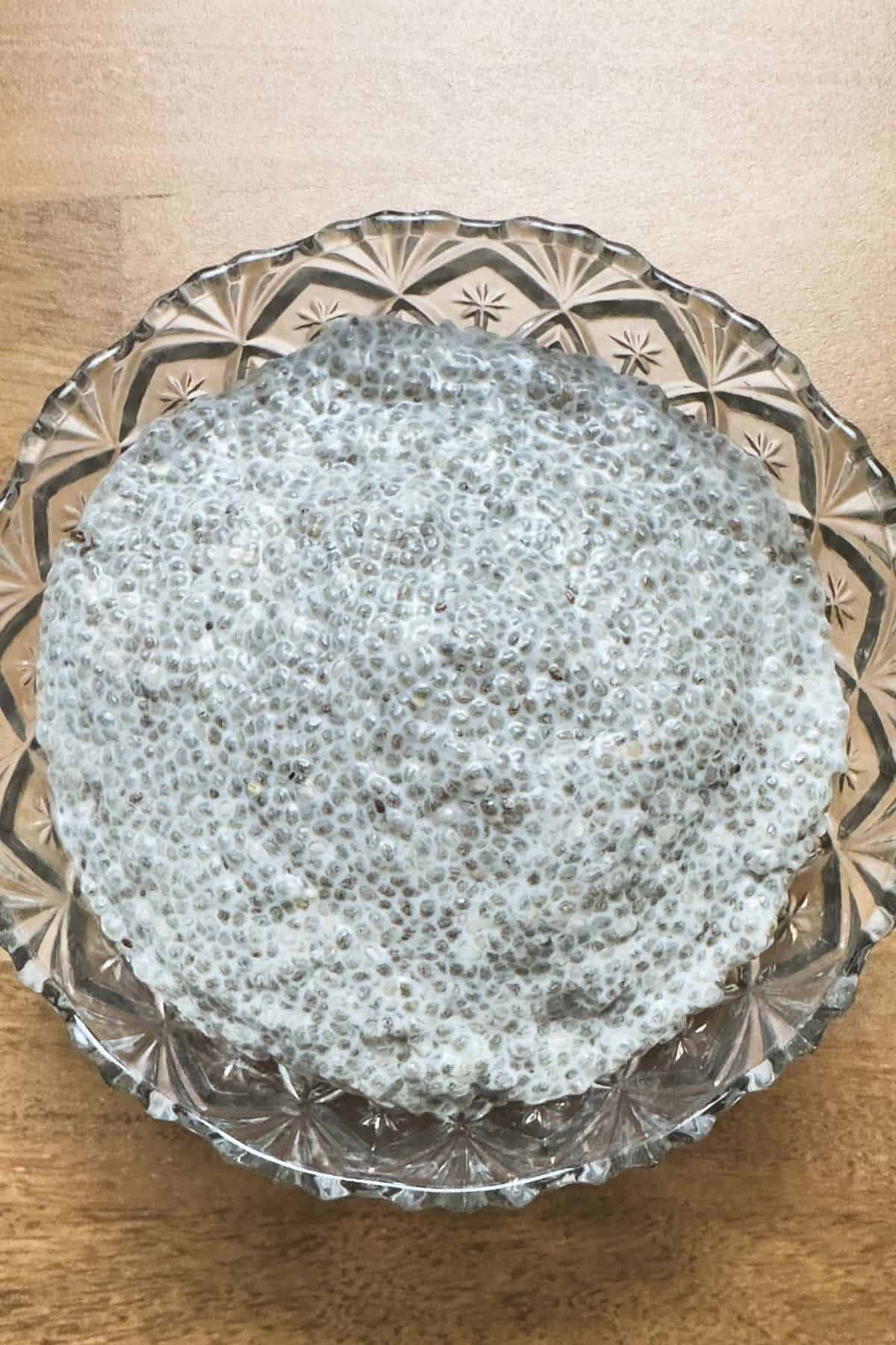 A glass bowl of mixed chia seeds and coconut milk on a wooden surface, with a spoon, showing the start of the gelling process for chia pudding after setting for 2 hours.