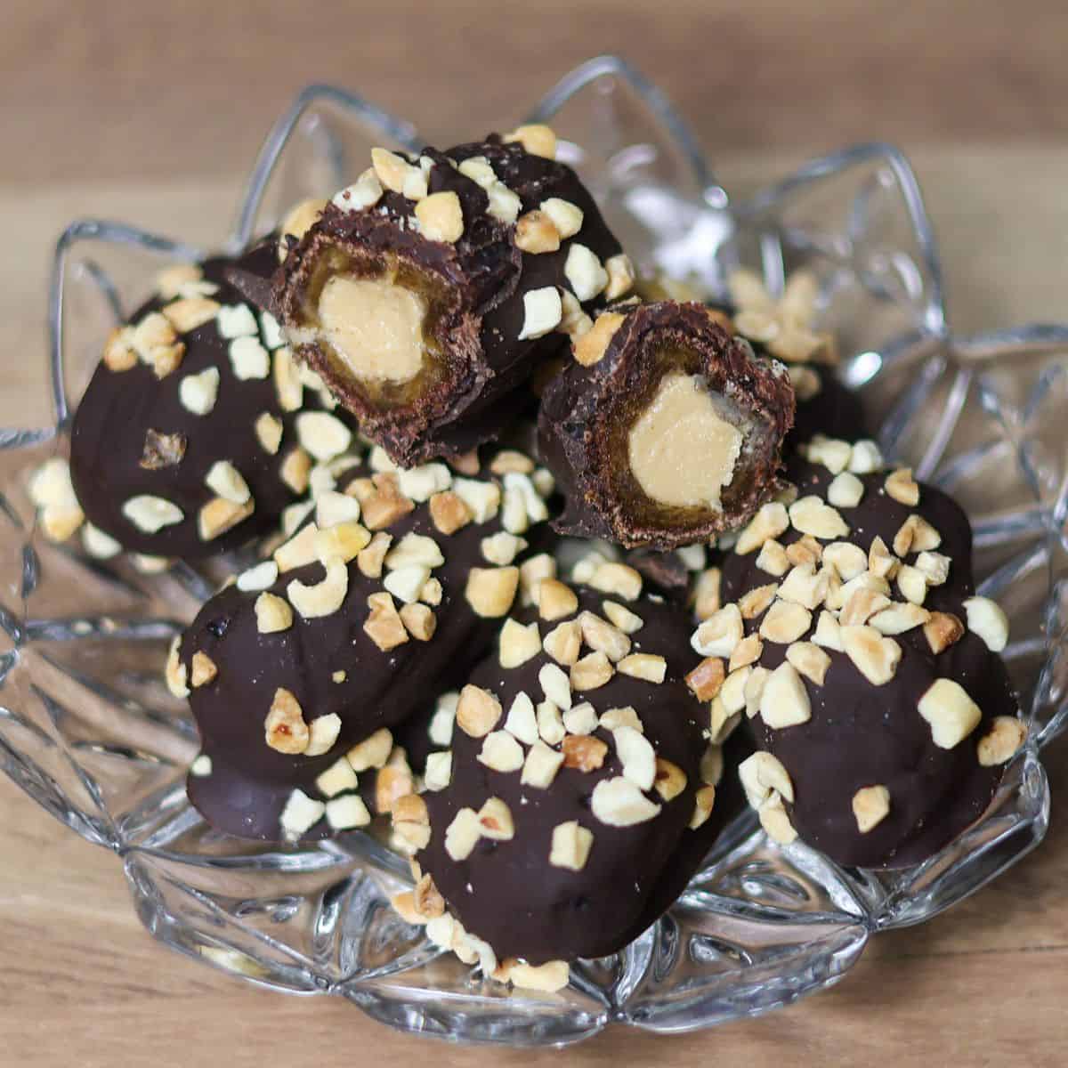Several chocolate covered dates with peanut butter filling and nut toppings arranged neatly on a glass dish.