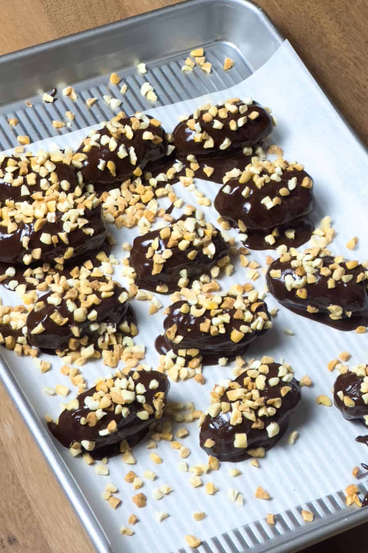 Freshly chocolate-covered stuffed dates generously topped with crushed nuts laid out on a baking sheet lined with parchment paper, ready to be chilled.