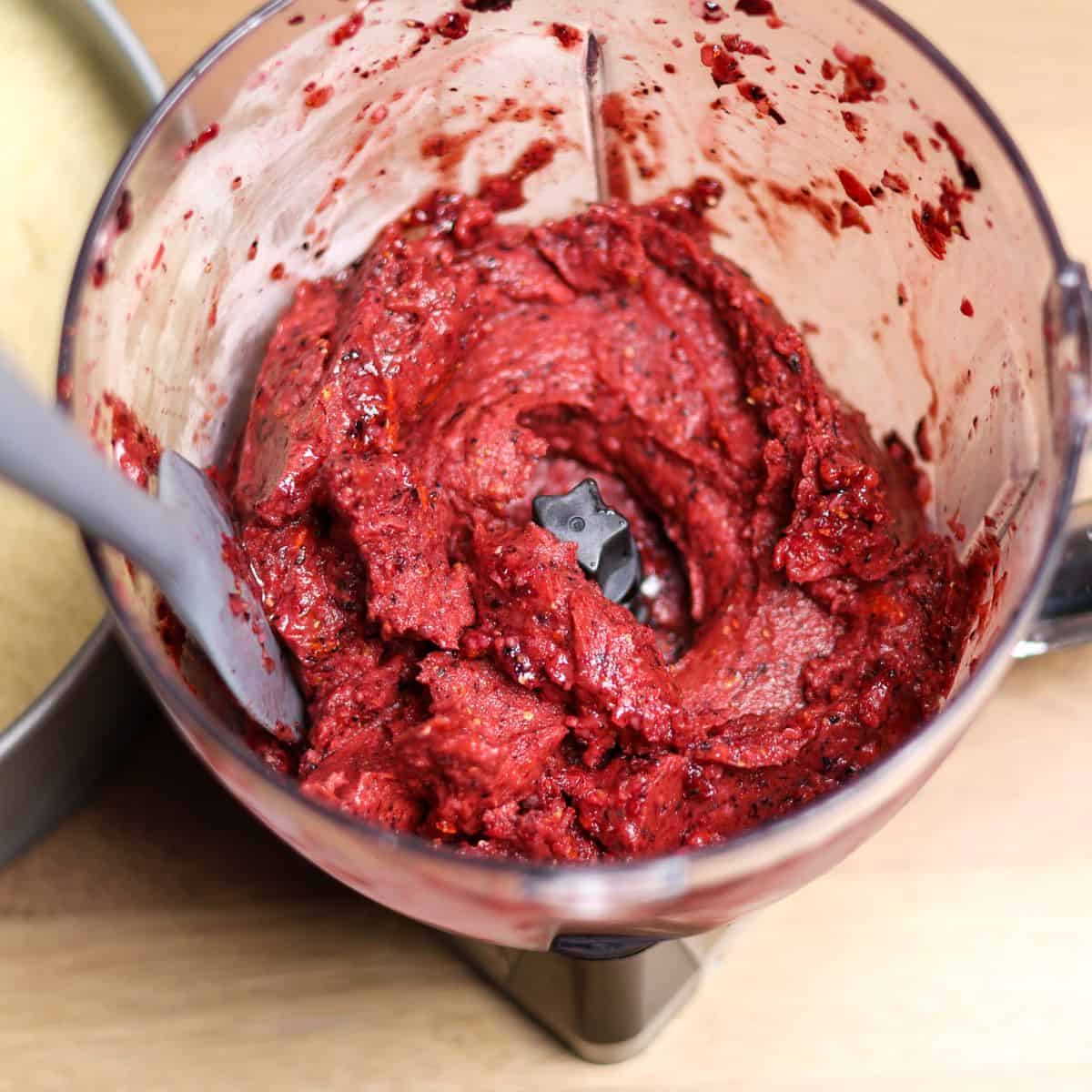 A blender filled with a rich, deep red berry mixture for a no-bake cheesecake topping, showing a swirled texture.