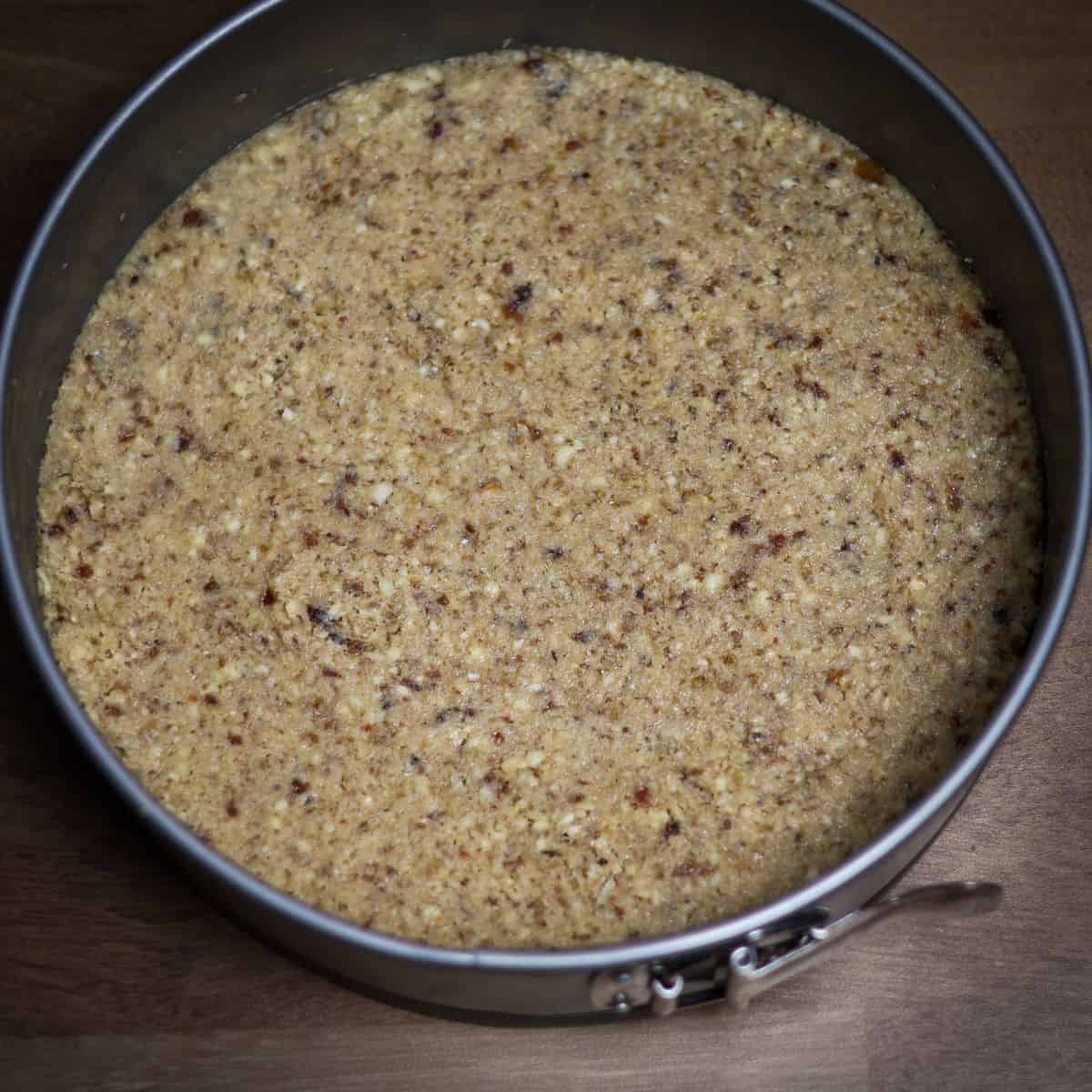 A round pan with a pressed crust of walnuts and dates, the base for a no-bake berry cheesecake, with a textured surface.