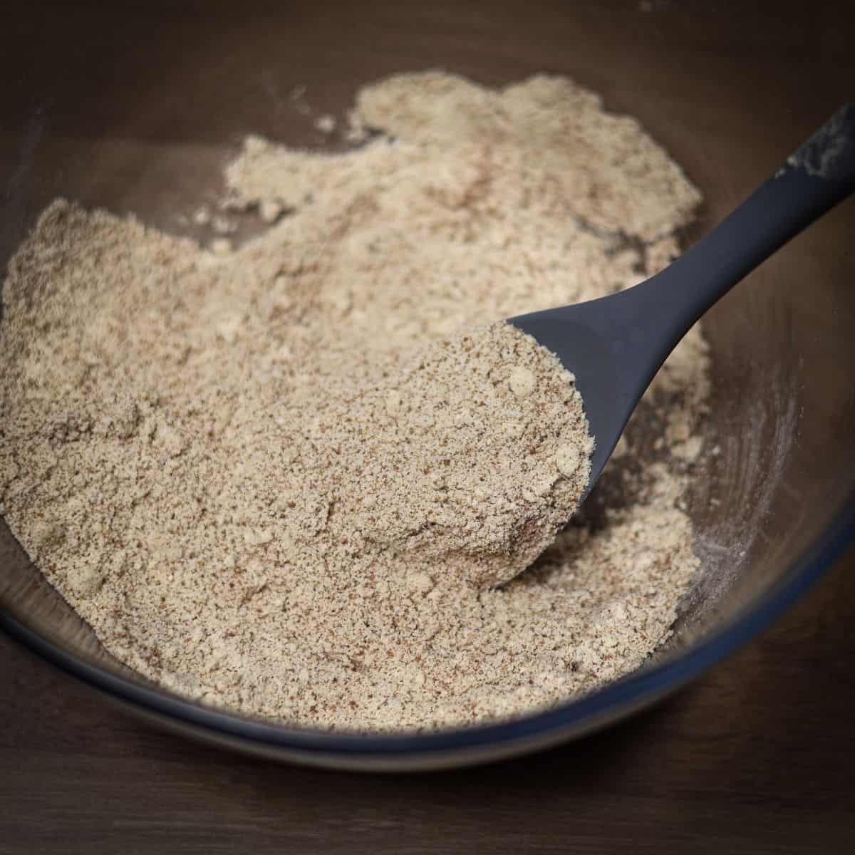 A bowl of dry ingredients for protein snickerdoodle cookies with a black silicone spatula. The mix appears finely ground and sandy in texture.