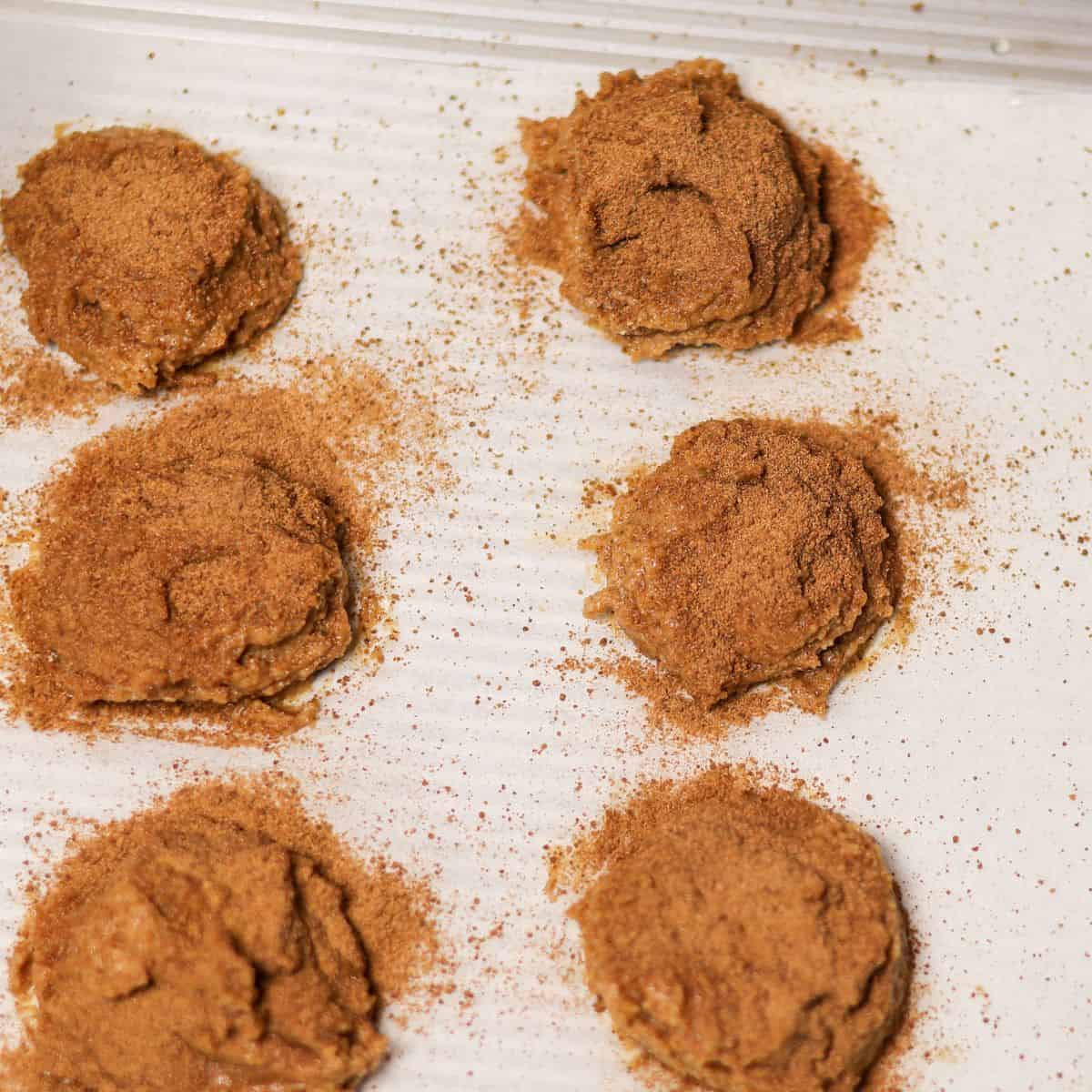 Portions of snickerdoodle cookie dough on a parchment-lined baking sheet, each sprinkled with a cinnamon and sugar mix.