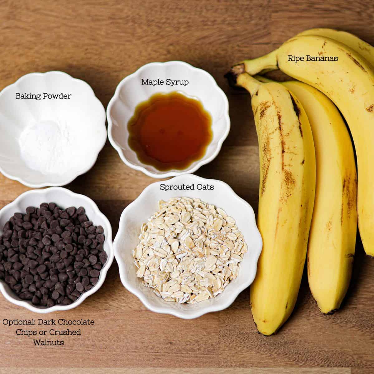 Ingredients for 4 Ingredient Sprouted Oat Banana Bread laid out on a wooden surface, including ripe bananas, sprouted oats, baking powder, and maple syrup, with optional dark chocolate chips or crushed walnuts.