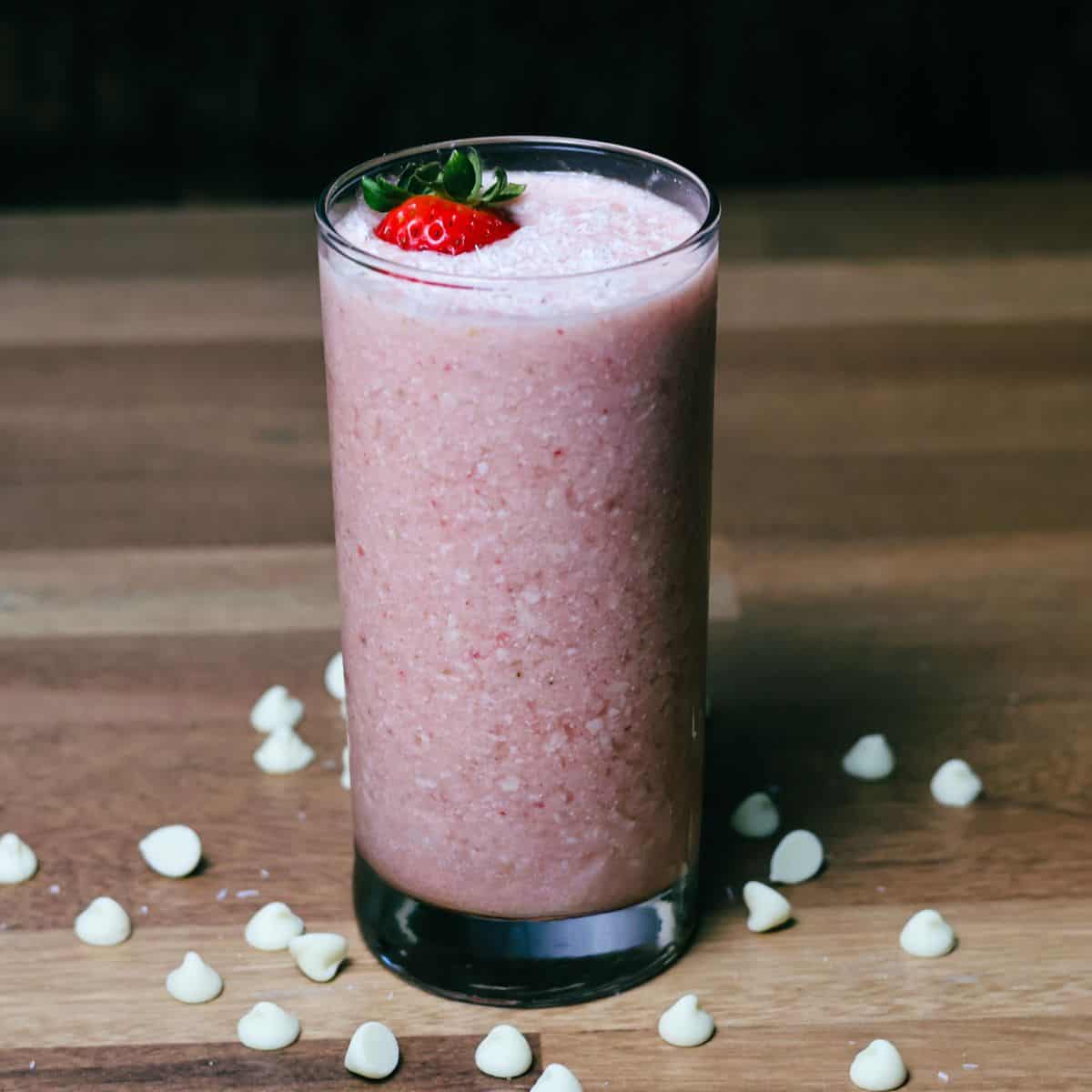 A tall, refreshing glass of pink Bahama Mama smoothie garnished with a strawberry, with white chocolate chips scattered on the wooden surface.