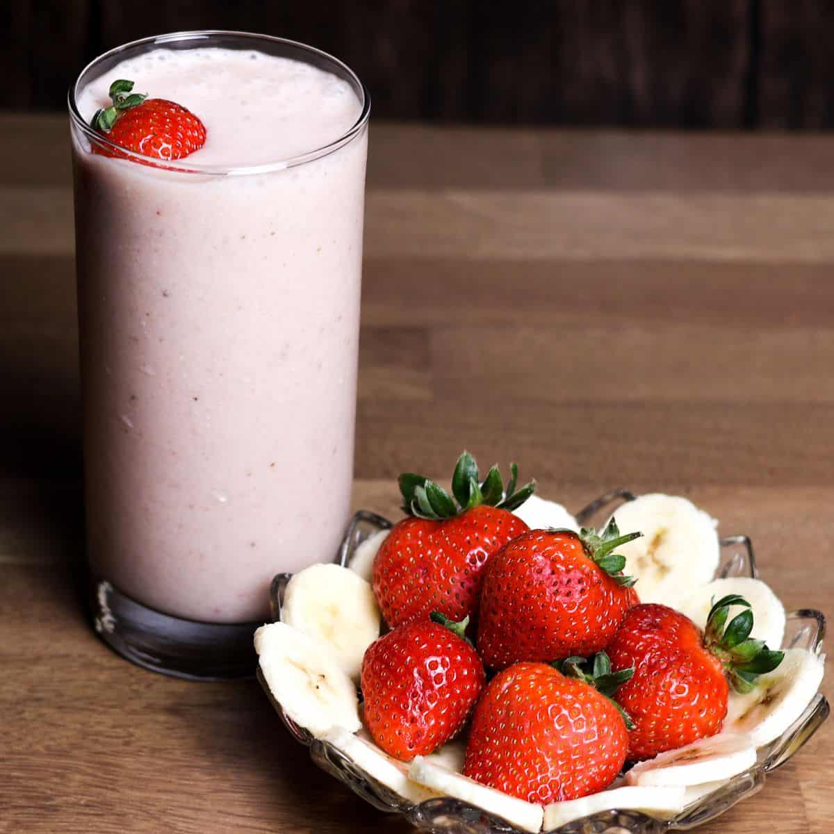 A side view of a tall glass filled with a smooth pink strawberry banana smoothie, beside a plate of whole strawberries and banana slices.