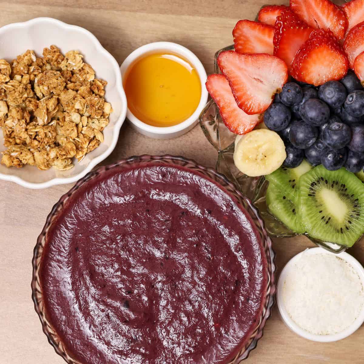 A bowl of blended açaí base ready for toppings, with separate bowls of cut fruits, granola, honey, and shredded coconut beside it.