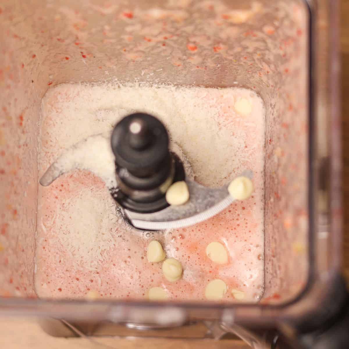 Overhead view of a blender with a creamy pink mixture topped with white chocolate chips and shredded coconut, indicating a midway point in a smoothie recipe