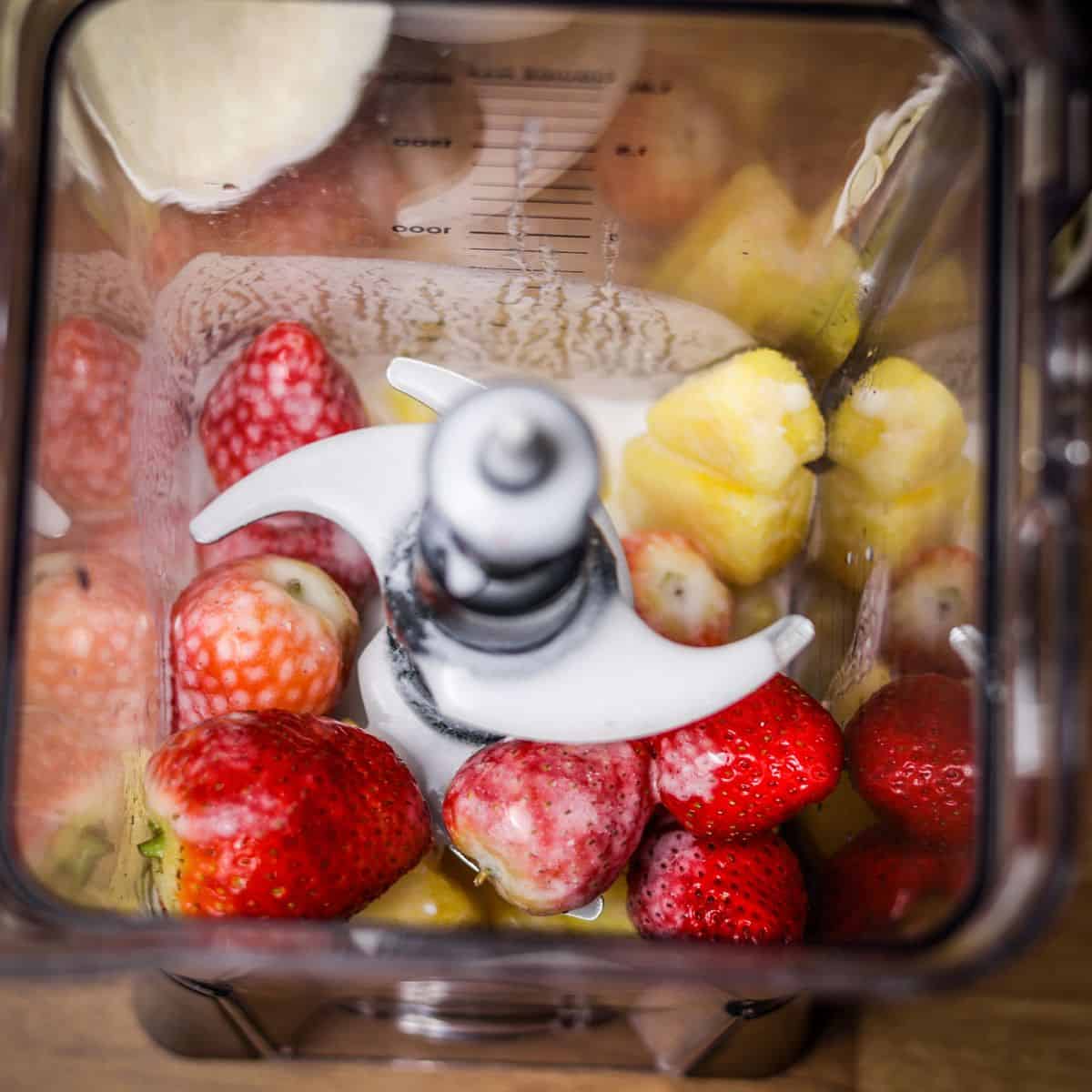 A blender from above filled with fresh whole strawberries and chunks of pineapple, pre-blend for a smoothie.