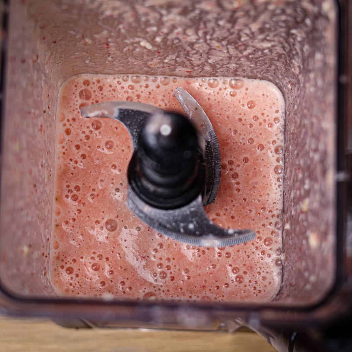 Looking directly down into a blender, showcasing a smooth pink strawberry pineapple smoothie after adding flavors.