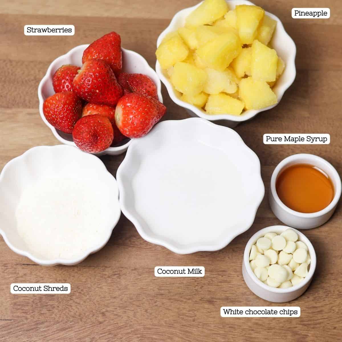 Neatly organized ingredients for a Bahama Mama smoothie, including bowls of strawberries, pineapple, white chocolate chips, shredded coconut, and maple syrup