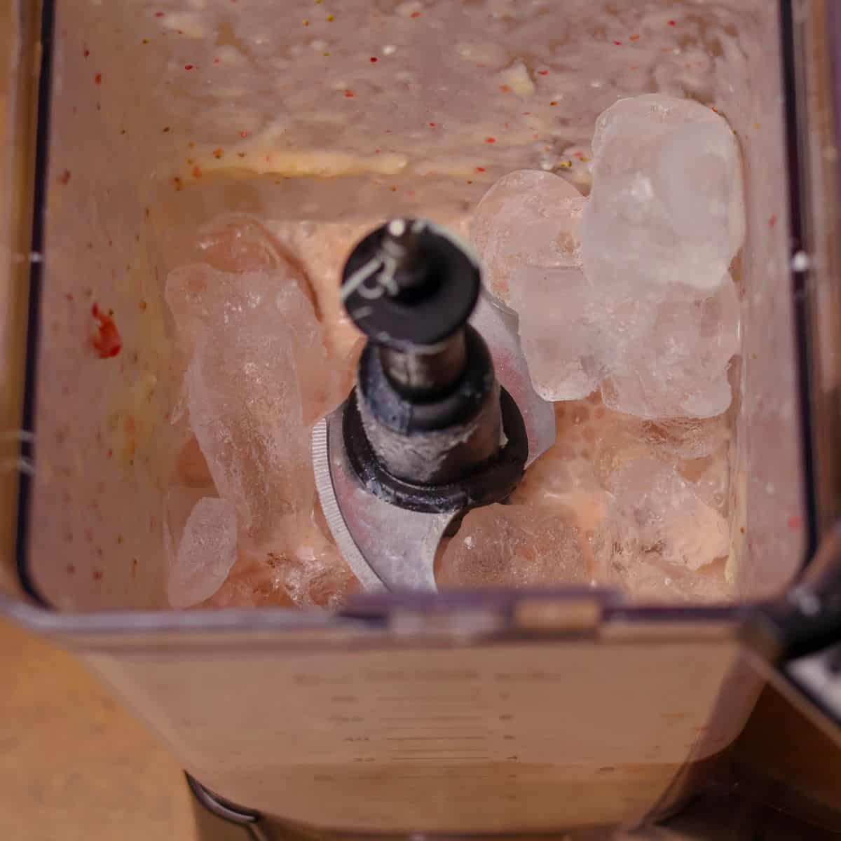 A blender filled with ice cubes on top of a partially blended mixture of pink smoothie, indicating a mid-blend pause to add ice for a frozen beverage.