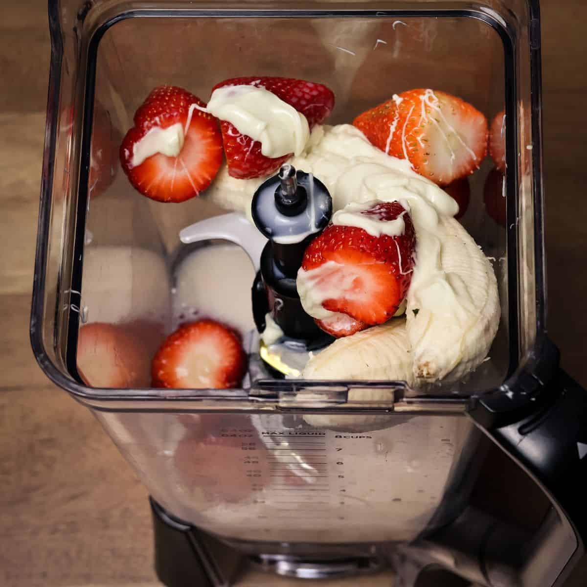 Top view of a blender with fresh strawberries, sliced bananas, and dollops of melted white chocolate on top, ready to be blended into a smoothie.