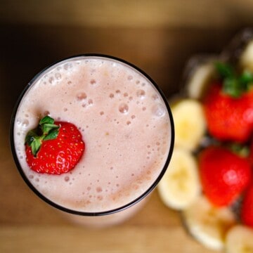 A bird's-eye view of a glass of pink smoothie topped with a whole strawberry, surrounded by more strawberries and banana slices on a wooden table.