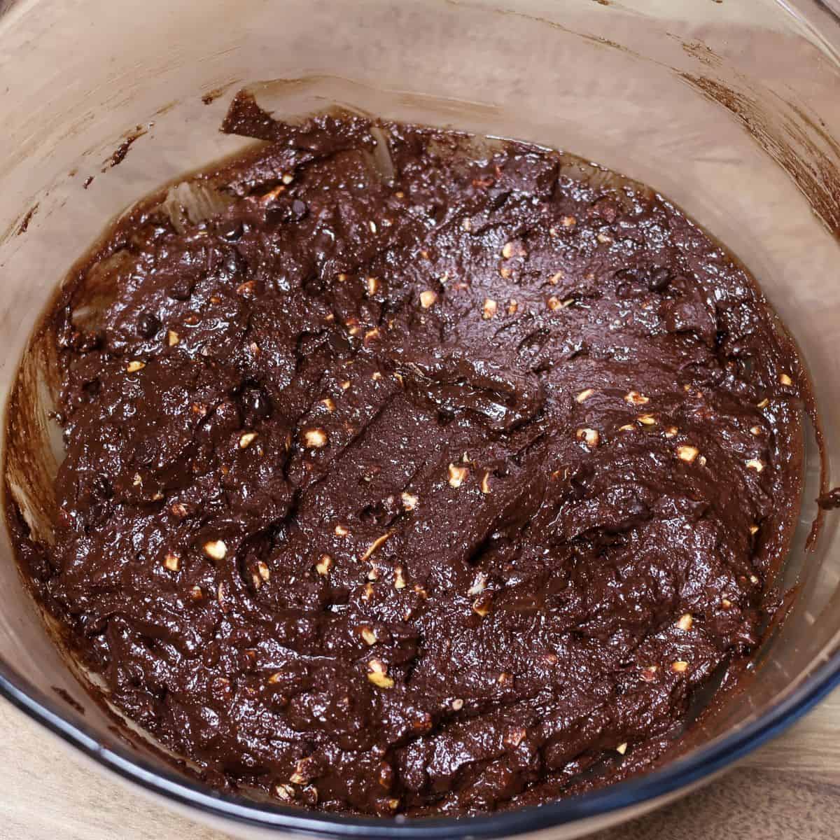 Thick, glossy brownie batter in a glass mixing bowl with nuts and chocolate chips folded in, ready for baking.