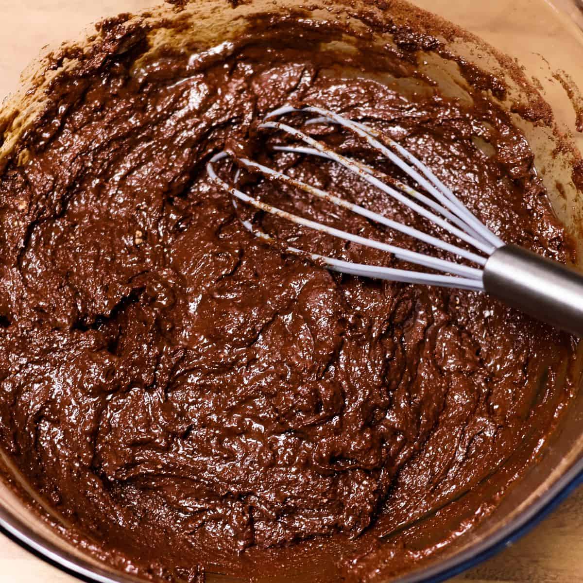 A whisk mixing the wet and dry ingredients together in a bowl to create a smooth, dark brownie batter.