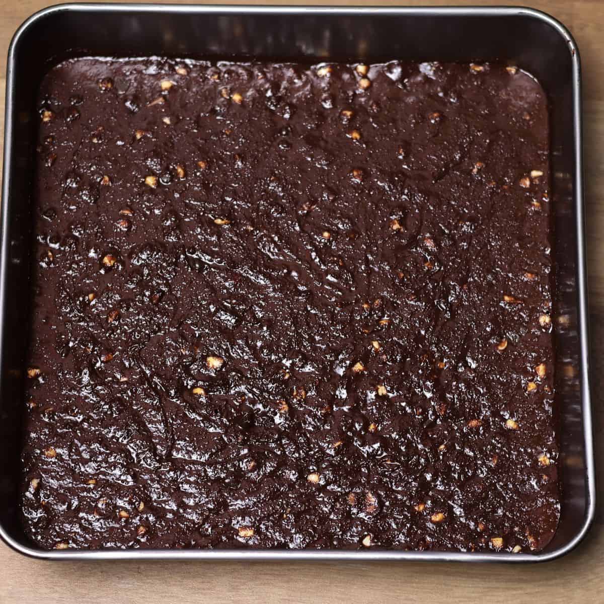 Brownie batter evenly spread in a square pan, speckled with nuts, ready to be baked in the oven.