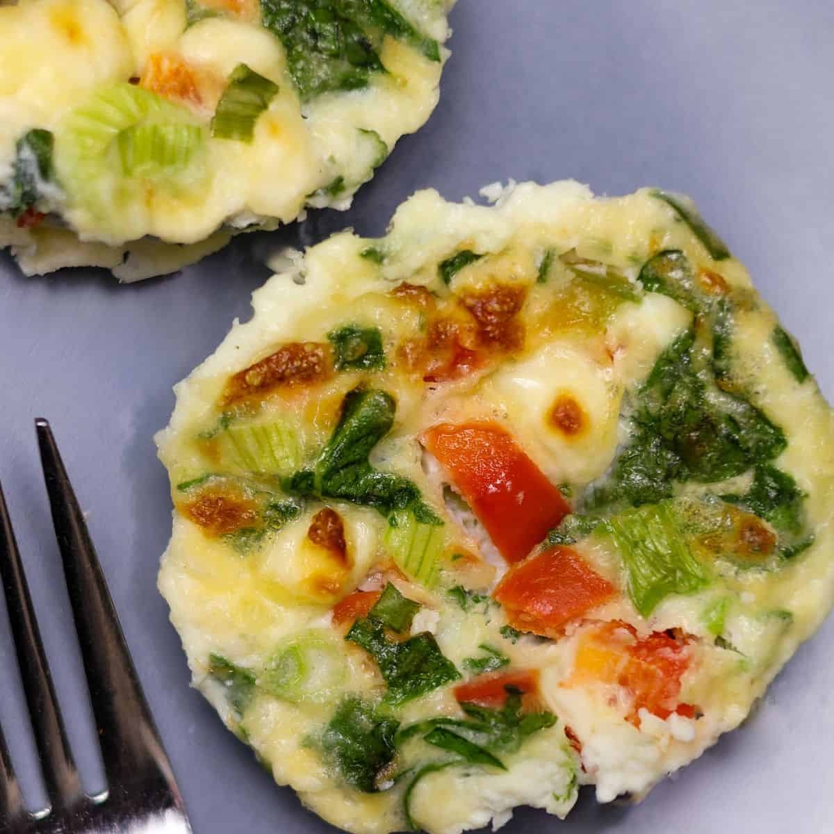Two egg white bites on a plate with visible chunks of green onion, red bell pepper, and spinach, with a silver fork on the side
