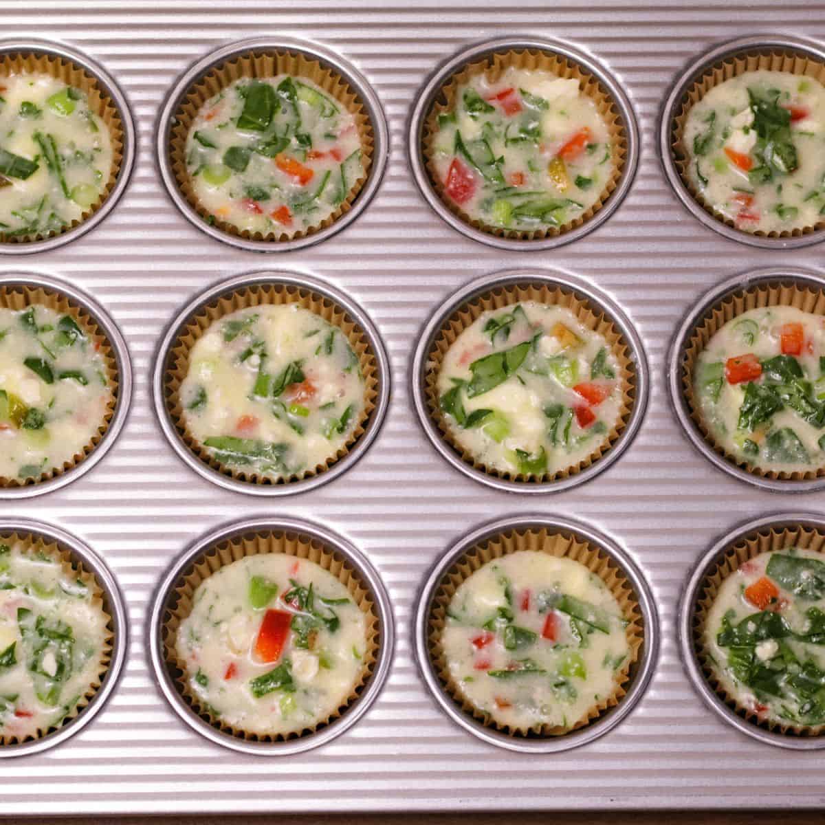 A muffin tin lined with paper liners filled with the raw egg white mixture, dotted with green and red from the spinach and bell peppers, ready to be baked.