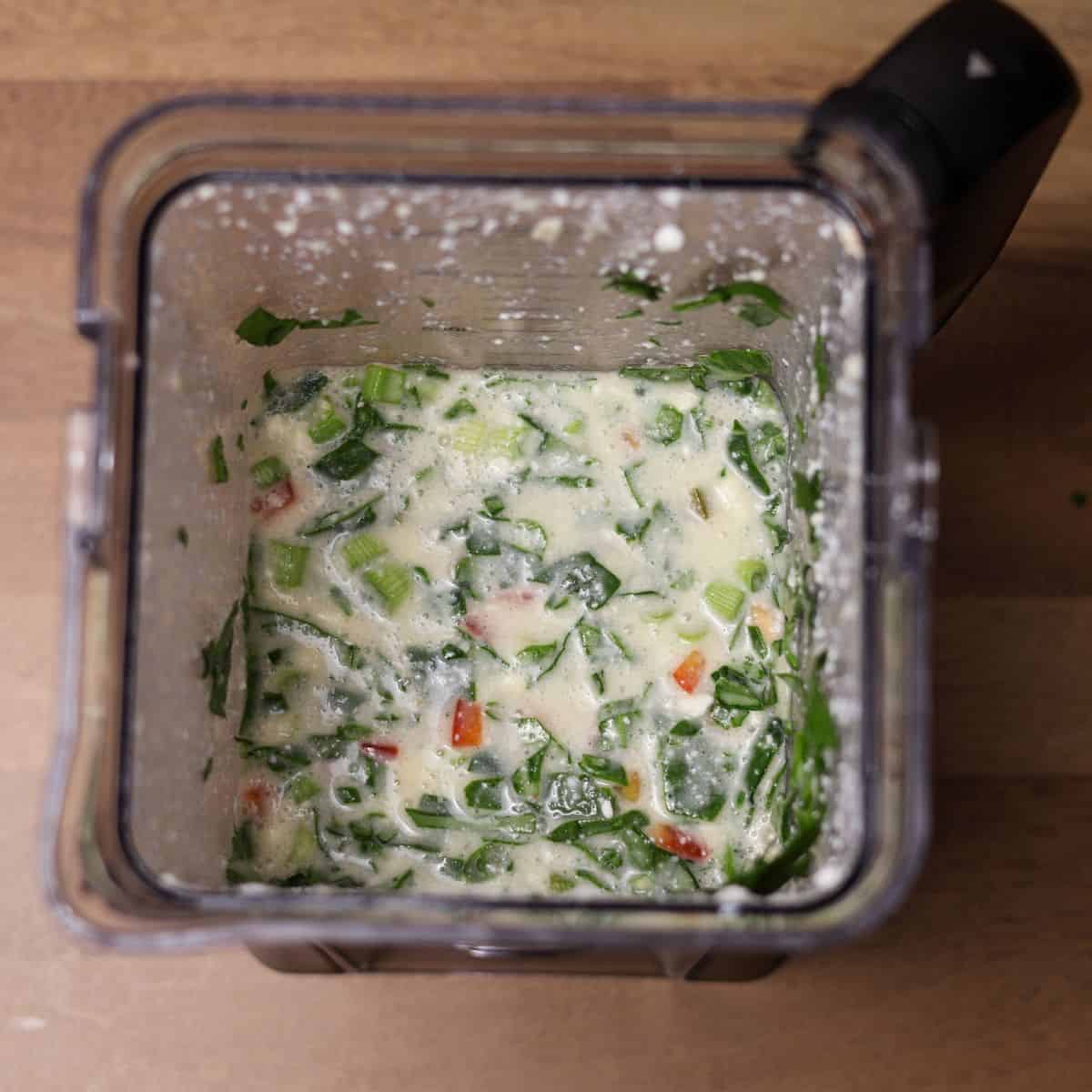 Looking down into a blender where the egg mixture is combined with finely chopped spinach, red bell peppers, and green onions, ready for the final mix.