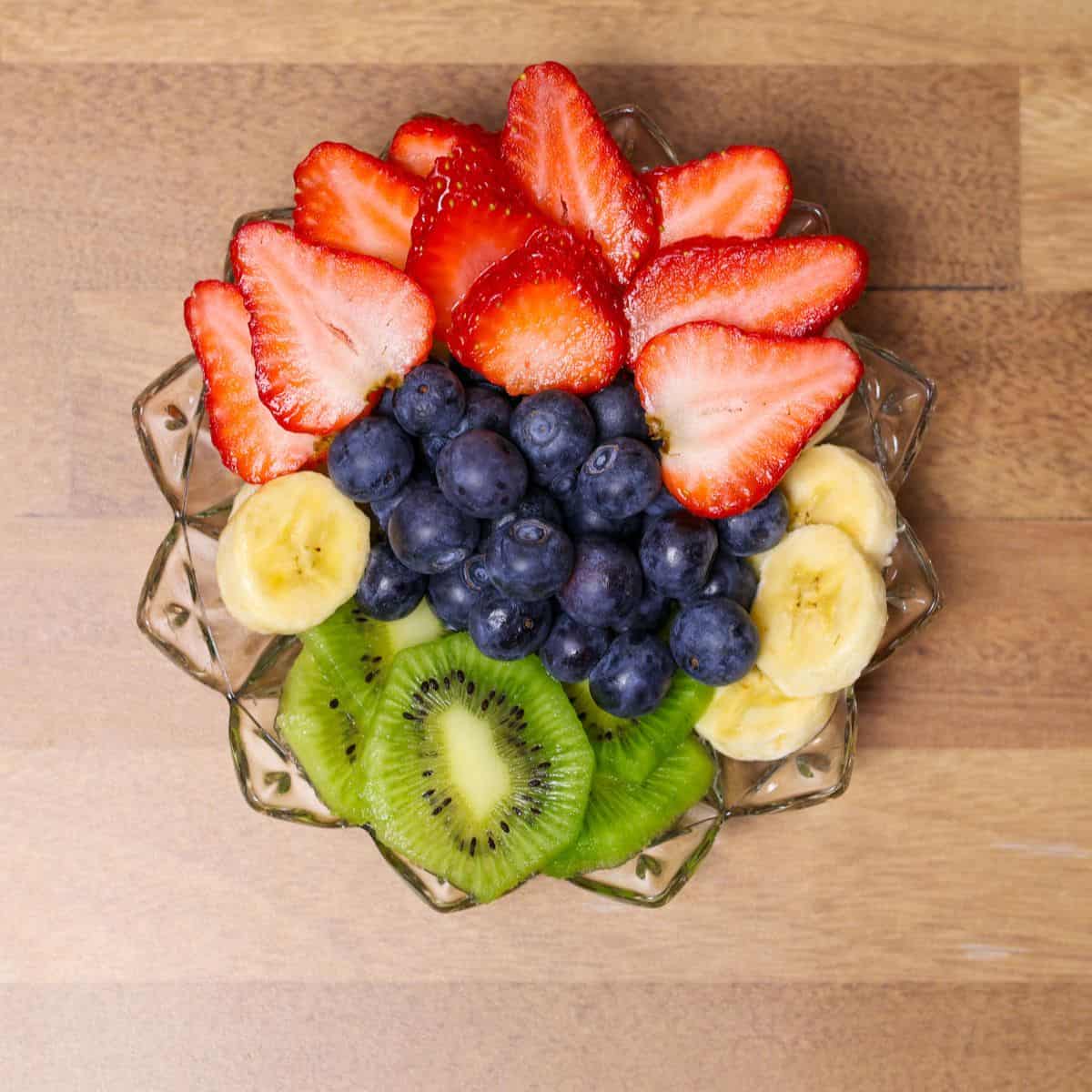An assortment of fresh fruits arranged on a plate, prepared as toppings for a Hawaiian açaí bowl, including sliced bananas, kiwi, strawberries, and whole blueberries