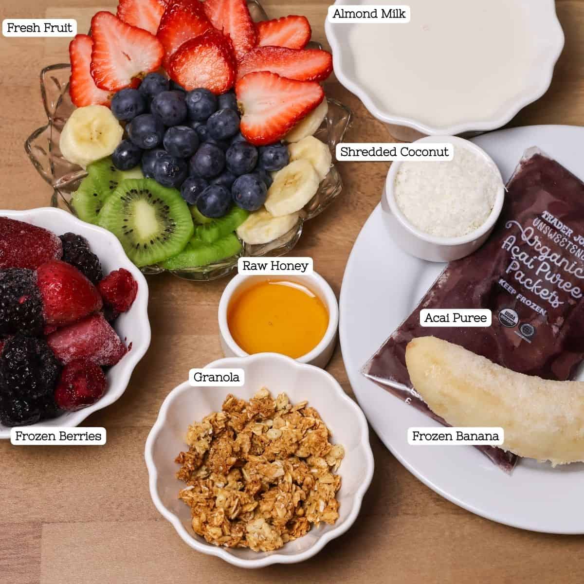 An overhead view of all the Hawaiian açaí bowl ingredients laid out on a table, including açaí puree, frozen berries, almond milk, granola, bananas, kiwi, strawberries, blueberries, shredded coconut, and raw honey.