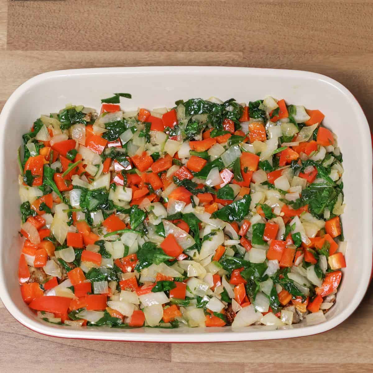 Chopped vegetables including red bell peppers, onions, and spinach layered on top of the meat in a white ceramic baking dish, prepped for a keto breakfast casserole.