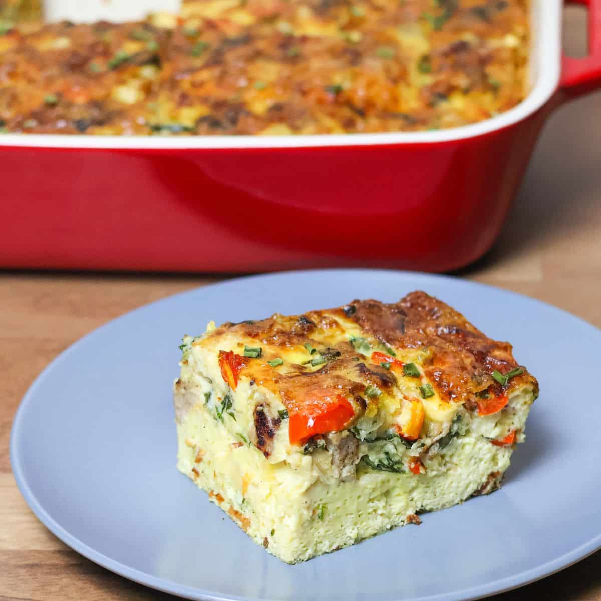 A full keto breakfast casserole in a red ceramic baking dish displayed on a wooden table, showing a top view of the baked, golden-brown crust with visible herbs.