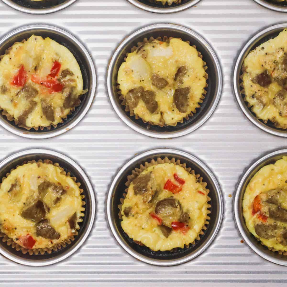 Golden brown keto egg bites fresh out of the oven in a muffin tin, showing the melted cheese and bits of red pepper and turkey sausage.