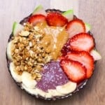 A completed acai bowl with peanut butter, granola, sliced bananas, strawberries, and green apple on top, presented in a decorative glass bowl.