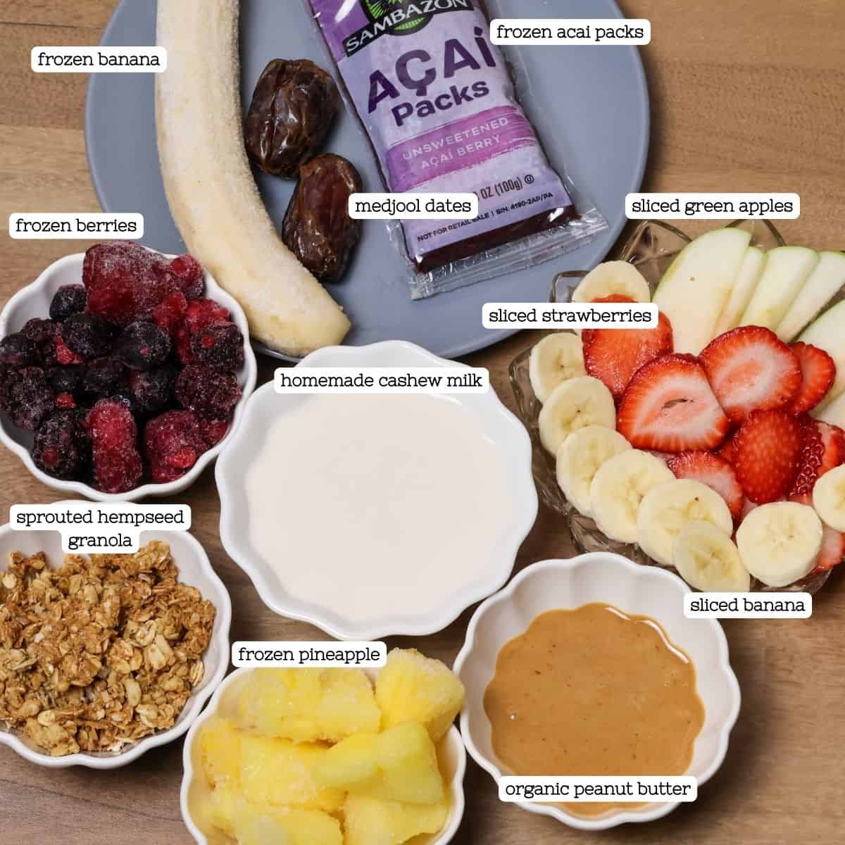 Laid out ingredients for an acai peanut butter bowl including frozen banana, berries, acai packs, green apples, strawberries, granola, frozen pineapple, cashew milk, and peanut butter.
